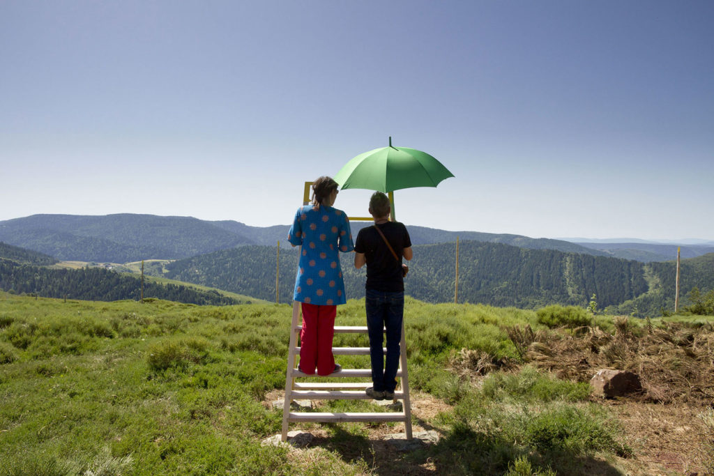 2 personnes sur une échelle observent un paysage. L'une tient un parapluie vert.